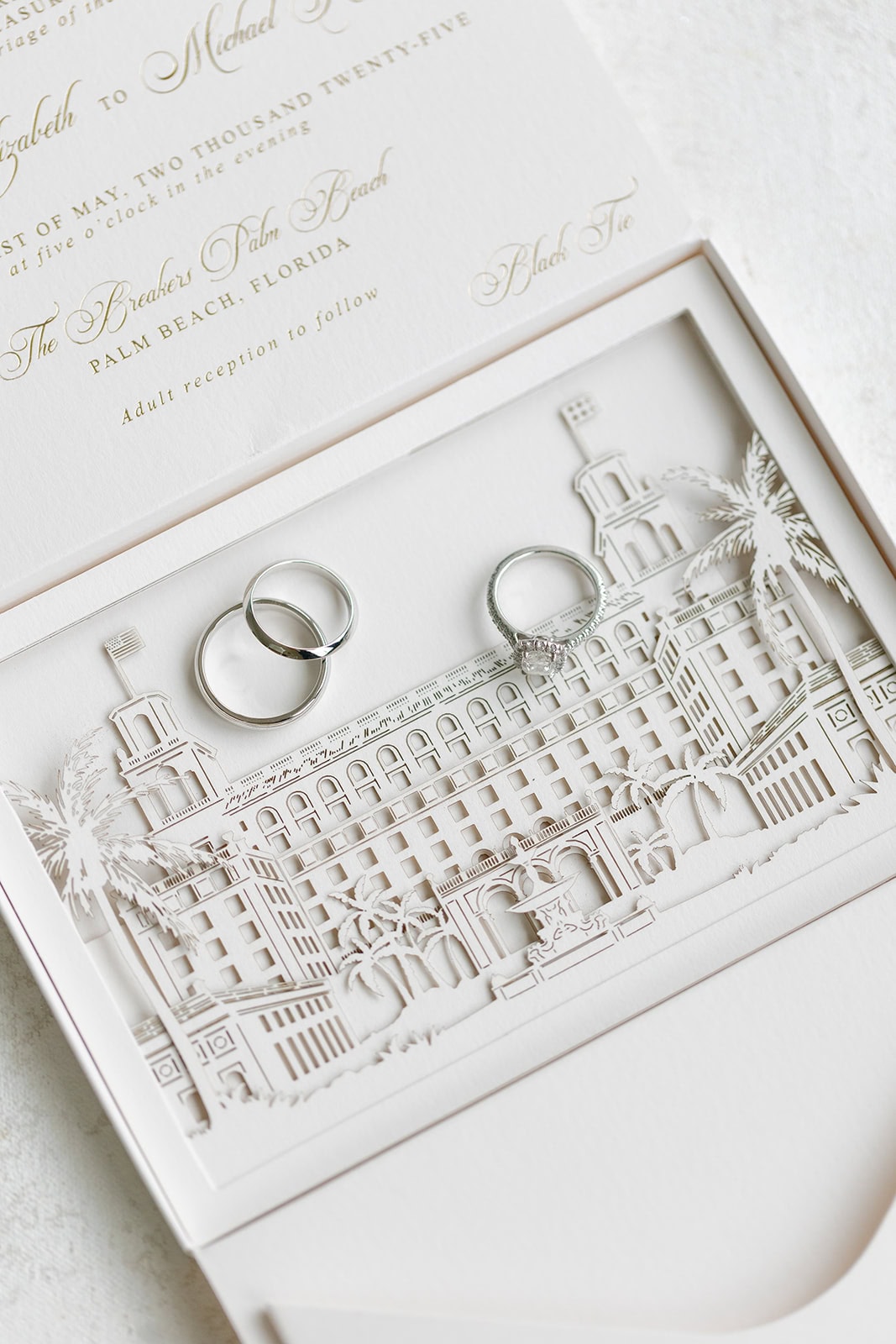 All white wedding invitation with wedding rings lying on top of the paper cut out our image of The Breakers with palm trees and fountain